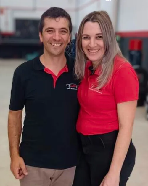 Um homem e uma mulher com o uniforme da Center Car, sorrindo dentro da oficina, com pneus ao fundo
